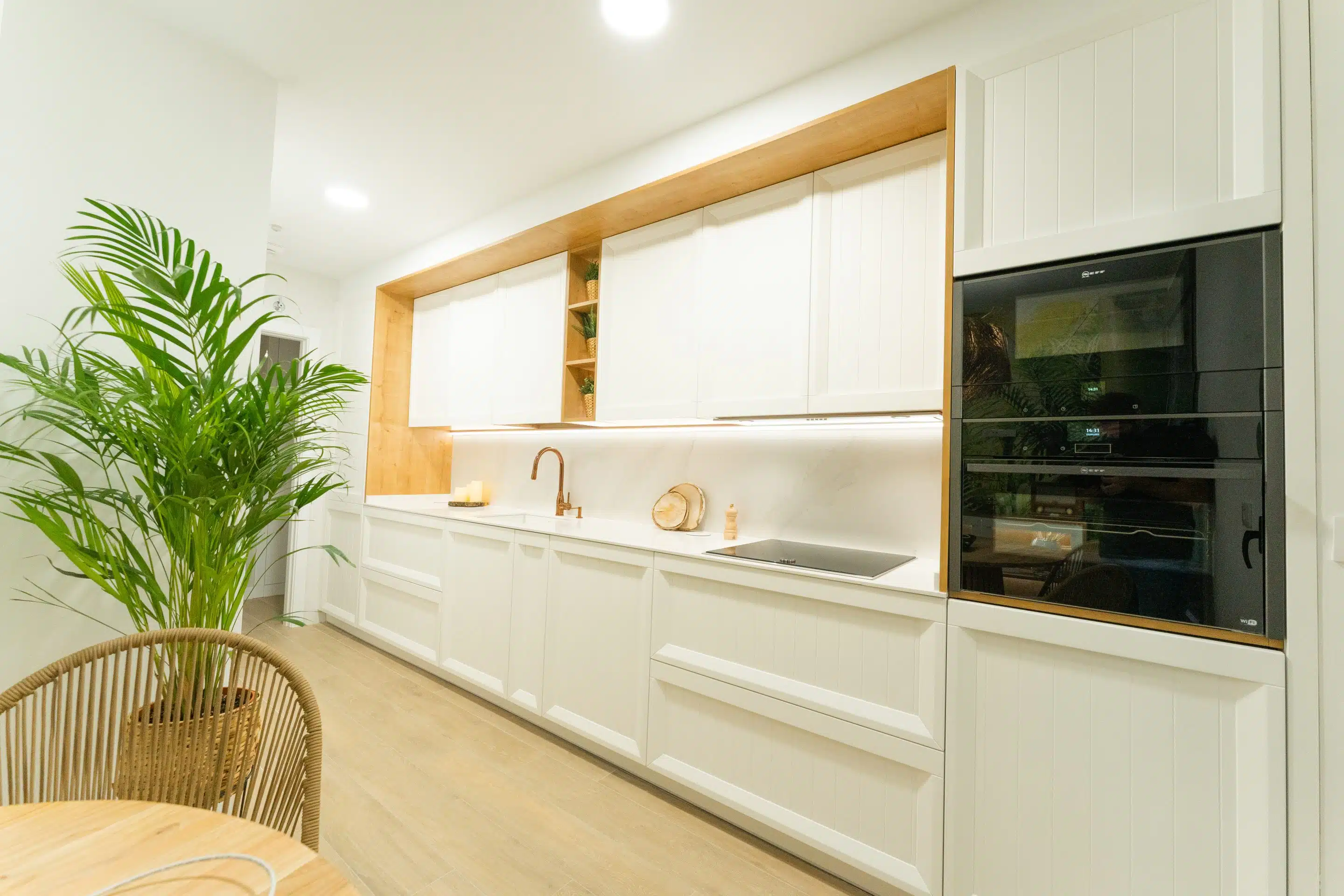 Vista lateral de esta cocina blanca clásica con estilos nórdicos donde la madera remarca el espacio y los hornos integrados rompen con el diseño para modernizar la composición