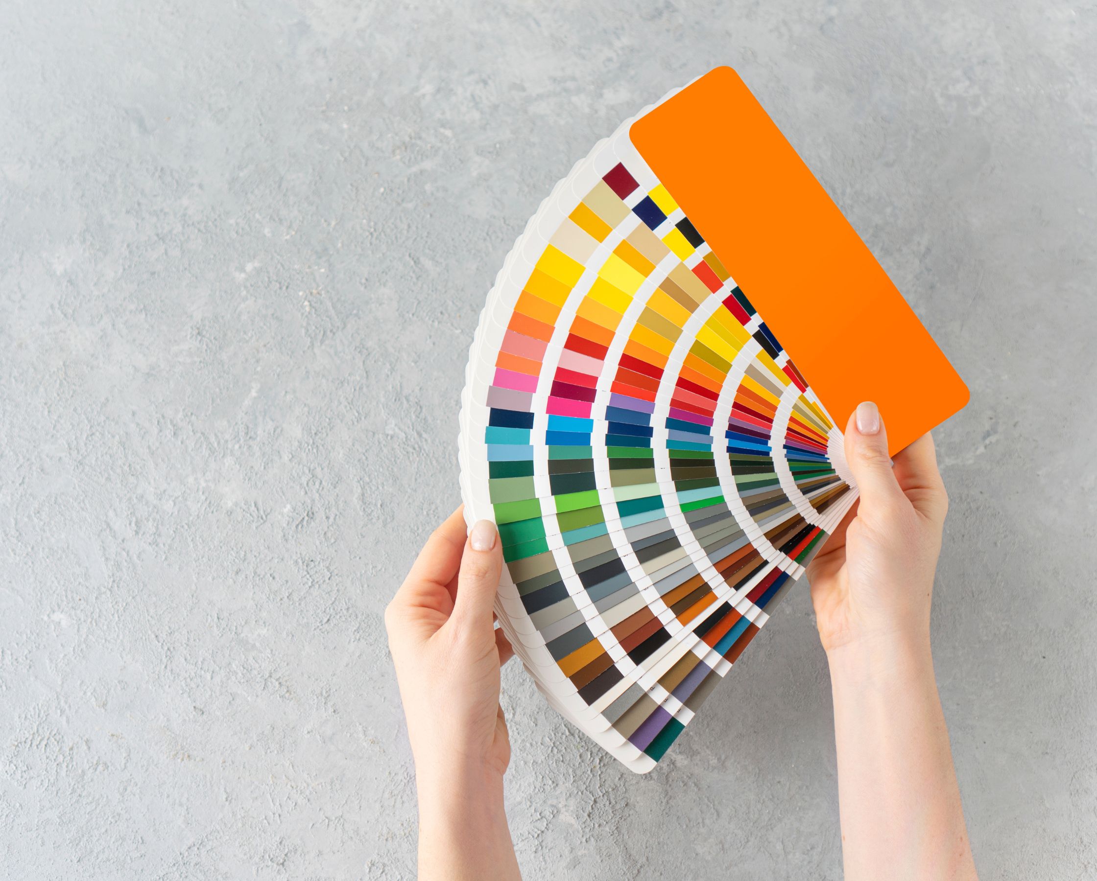 Una foto de dos manos sosteniendo un abanico de muestras de colores de pintura, desplegado para mostrar una amplia gama de colores que incluye tonos de naranja, amarillo, verde, azul, morado, marrón y gris, con un fondo texturizado de concreto gris claro