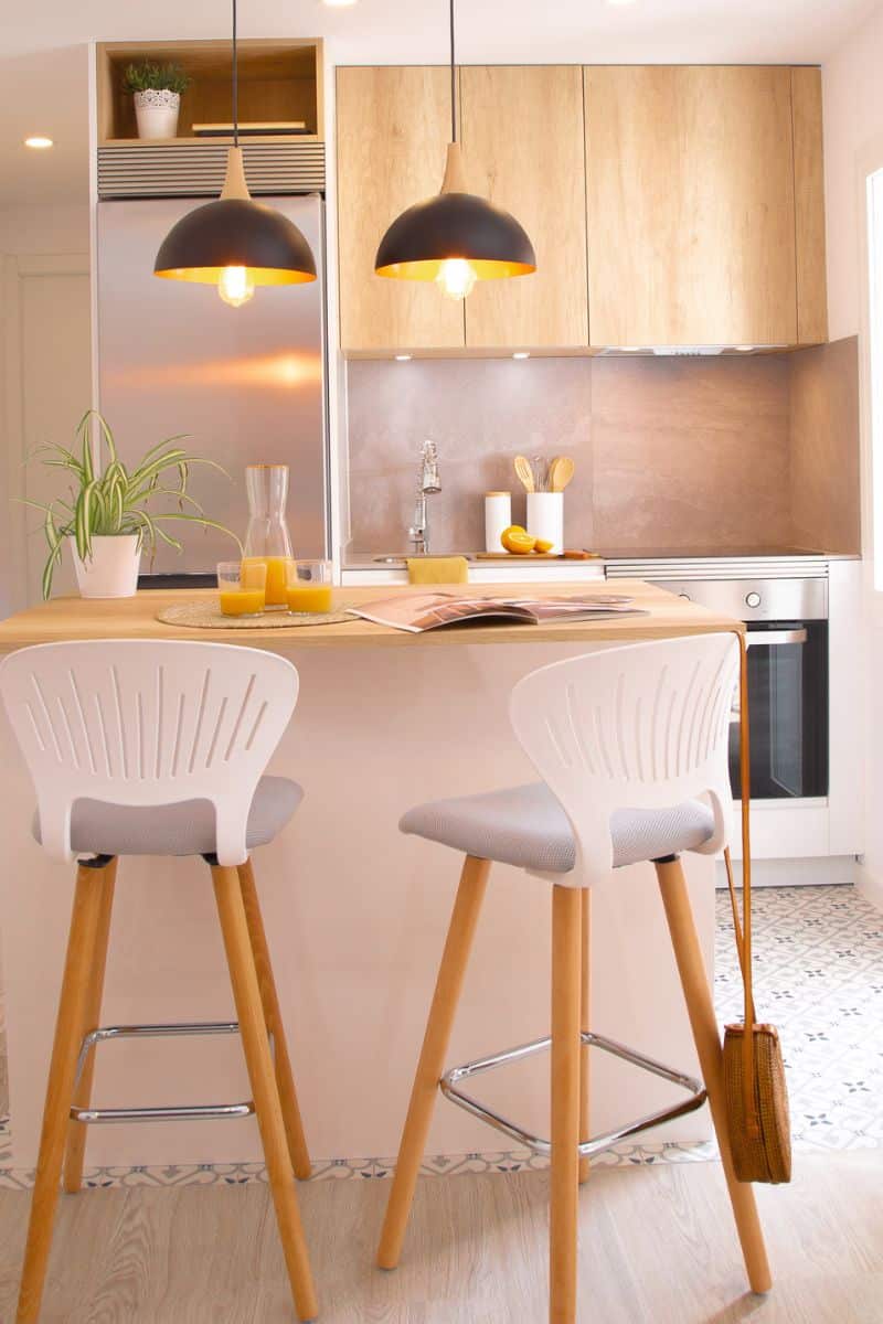 Foto de una cocina con una isla central de color claro, decorada con dos sillas altas de respaldo blanco y asiento gris, patas de madera y reposapiés metálico. Encima de la isla hay dos vasos de jugo de naranja, una jarra y una revista abierta. Dos lámparas negras de techo cuelgan sobre la isla. Al fondo, una encimera con grifo metálico, utensilios de cocina en recipientes blancos y un horno empotrado. A la izquierda, un refrigerador de acero inoxidable y una planta en maceta pequeña.