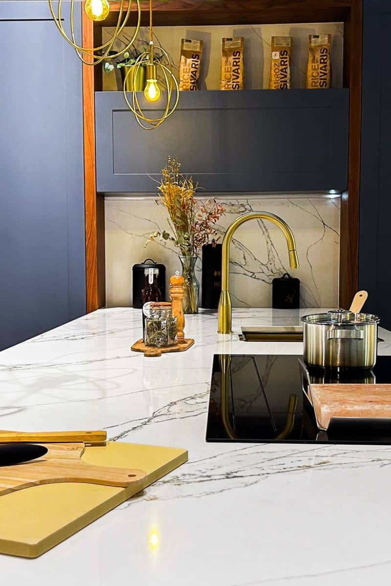 Foto de una cocina moderna con una encimera de mármol blanco con vetas grises. Sobre la encimera hay una tabla de cortar de madera, un frasco de hierbas, un molinillo de pimienta, un florero con plantas secas y una olla metálica en una estufa de inducción. Al fondo, un grifo dorado curvo y varios recipientes de almacenamiento, incluyendo uno negro para pasta. Encima, se ven bolsas de arroz Sivaris en una repisa. Dos lámparas colgantes con jaulas doradas rodean las bombillas expuestas.
