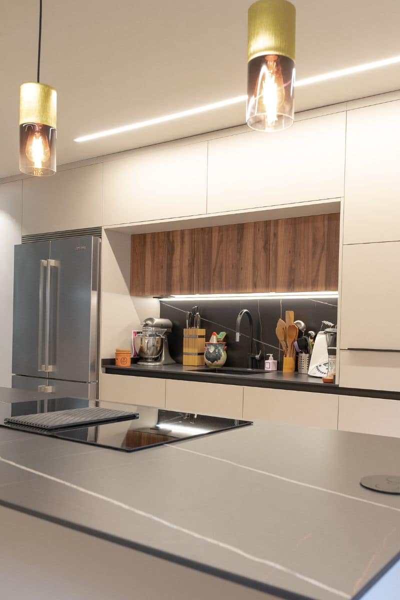 Foto de una cocina moderna con una gran isla central gris y dos lámparas colgantes de vidrio y metal que cuelgan del techo. La encimera incluye una estufa integrada de inducción. Al fondo, una encimera negra con fregadero metálico, una batidora de pie, utensilios de cocina en un recipiente de madera y varios electrodomésticos. Los armarios superiores tienen un acabado de madera oscura, mientras que los inferiores y laterales son de color claro. A la izquierda, hay un refrigerador de acero inoxidable de dos puertas.