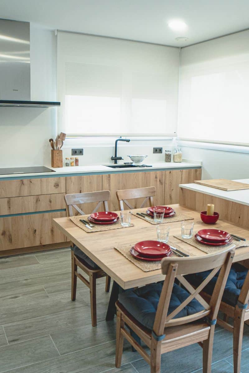 Foto de una cocina con una mesa de madera puesta para cuatro personas, con platos rojos, vasos de cristal y cubiertos colocados sobre manteles individuales de yute. Las sillas de madera tienen cojines grises. Al fondo, una encimera blanca con fregadero negro, colador de metal y varios utensilios de cocina. Los muebles son de madera clara, y las ventanas están cubiertas con persianas enrollables blancas que permiten la entrada de luz natural. A la izquierda, hay una estufa integrada con campana extractora de acero inoxidable.
