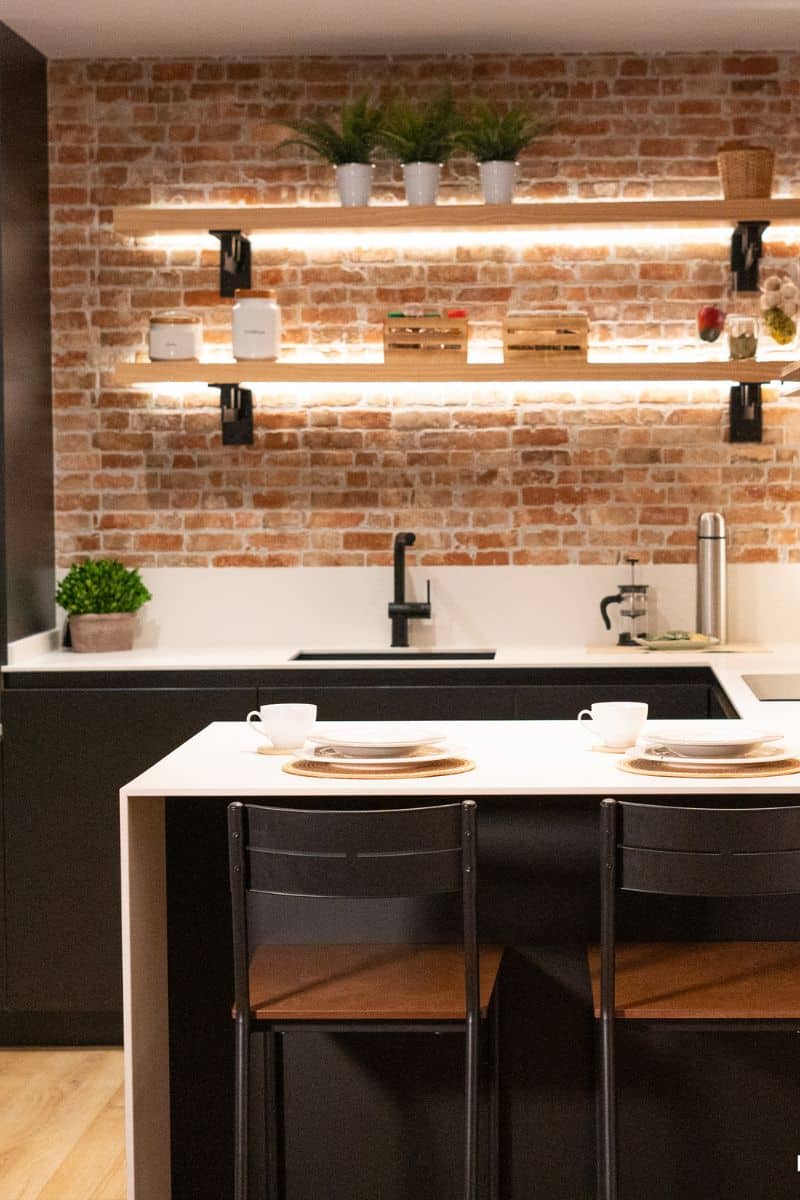 Foto de una cocina con una pared de ladrillo expuesto, equipada con estantes flotantes de madera iluminados, decorados con plantas en macetas blancas, tarros de almacenamiento y una caja de madera pequeña. La encimera es blanca con un fregadero y grifo negros. Al frente, hay una barra desayunadora con dos sillas negras de respaldo metálico, y está puesta para dos personas con platos blancos sobre manteles individuales redondos. A la derecha, hay una cafetera de prensa francesa y un termo plateado.